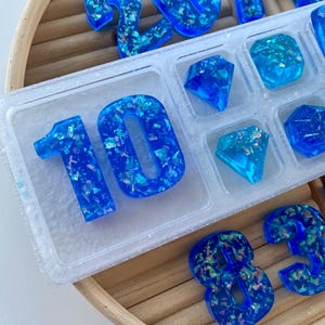 May include: Blue resin numbers and gem shapes with iridescent glitter are displayed in a clear plastic tray. The numbers include 10, 2, 3, 7, 4, and 8. The gem shapes are diamonds and squares. The tray sits on a wooden tray.