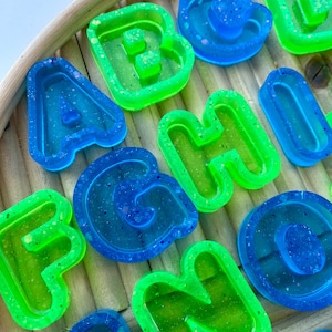 May include: Close-up of colourful resin letters in blue and green. The letters are arranged on a light brown, woven surface. The letters are in various fonts and sizes, with a glittery, translucent appearance.