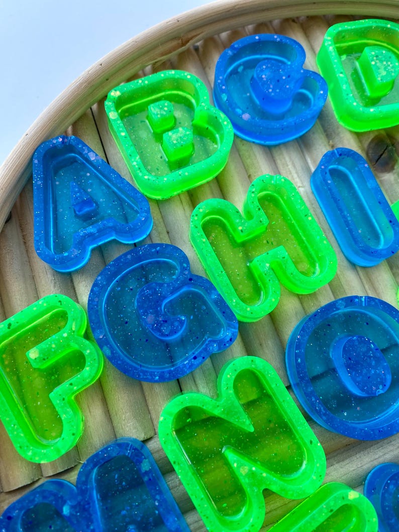 May include: Close-up of colourful resin letters in blue and green. The letters are arranged on a light brown, woven surface. The letters are in various fonts and sizes, with a glittery, translucent appearance.