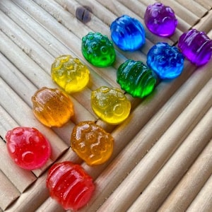 May include: An assortment of colorful, translucent, oval-shaped candies arranged on a bamboo tray. The candies are in shades of red, orange, yellow, green, blue, and purple. Each candy has a unique texture and design, creating a visually appealing display.