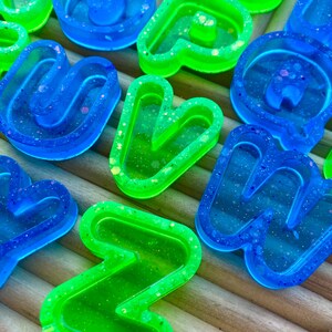 May include: Close-up of several resin letters in vibrant blue and green hues. The letters are filled with glitter and are arranged on a light brown, textured surface. The letters are in various shapes and sizes.