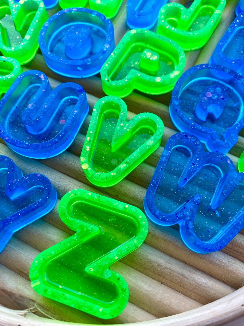 May include: Close-up of several resin letters in vibrant blue and green hues. The letters are filled with glitter and are arranged on a light brown, textured surface. The letters are in various shapes and sizes.