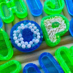 May include: A selection of resin art pieces in bright neon green and blue. Some are filled with white beads or rice, others are empty. The pieces are arranged on a wooden surface. Measurements are not applicable.