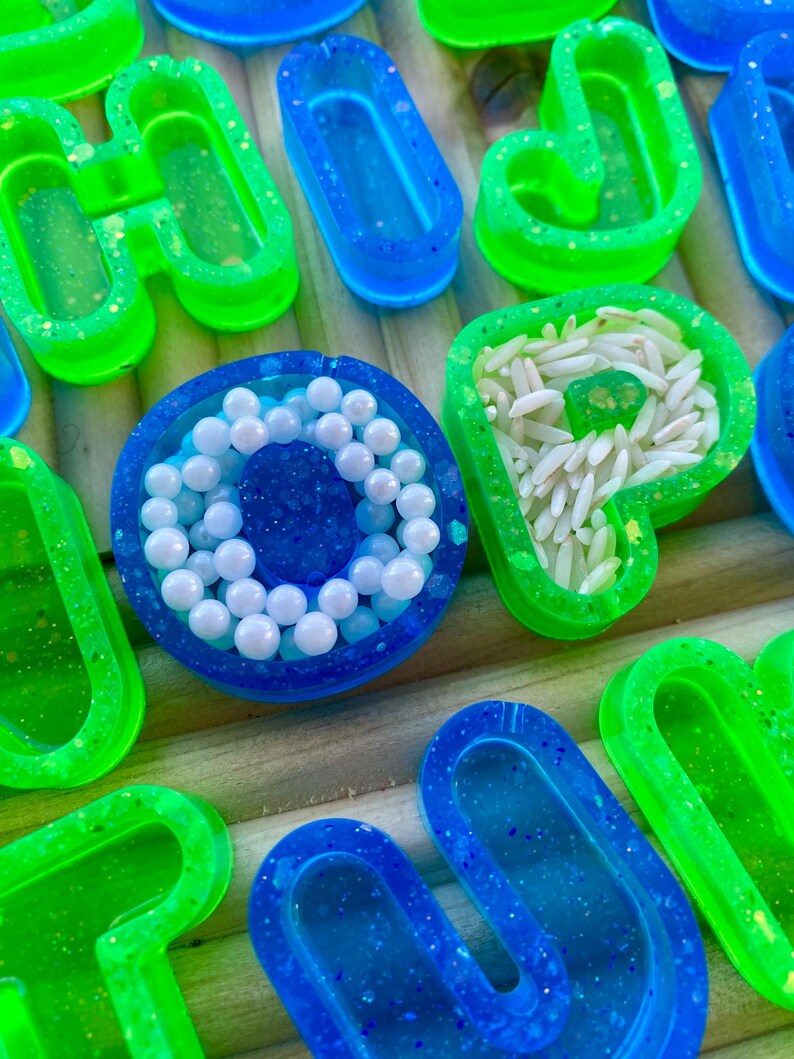 May include: A selection of resin art pieces in bright neon green and blue. Some are filled with white beads or rice, others are empty. The pieces are arranged on a wooden surface. Measurements are not applicable.
