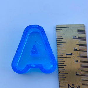 May include: A blue resin letter "A" with a glittery texture, next to a ruler. The letter is approximately 5 cm tall. The background is a light blue.