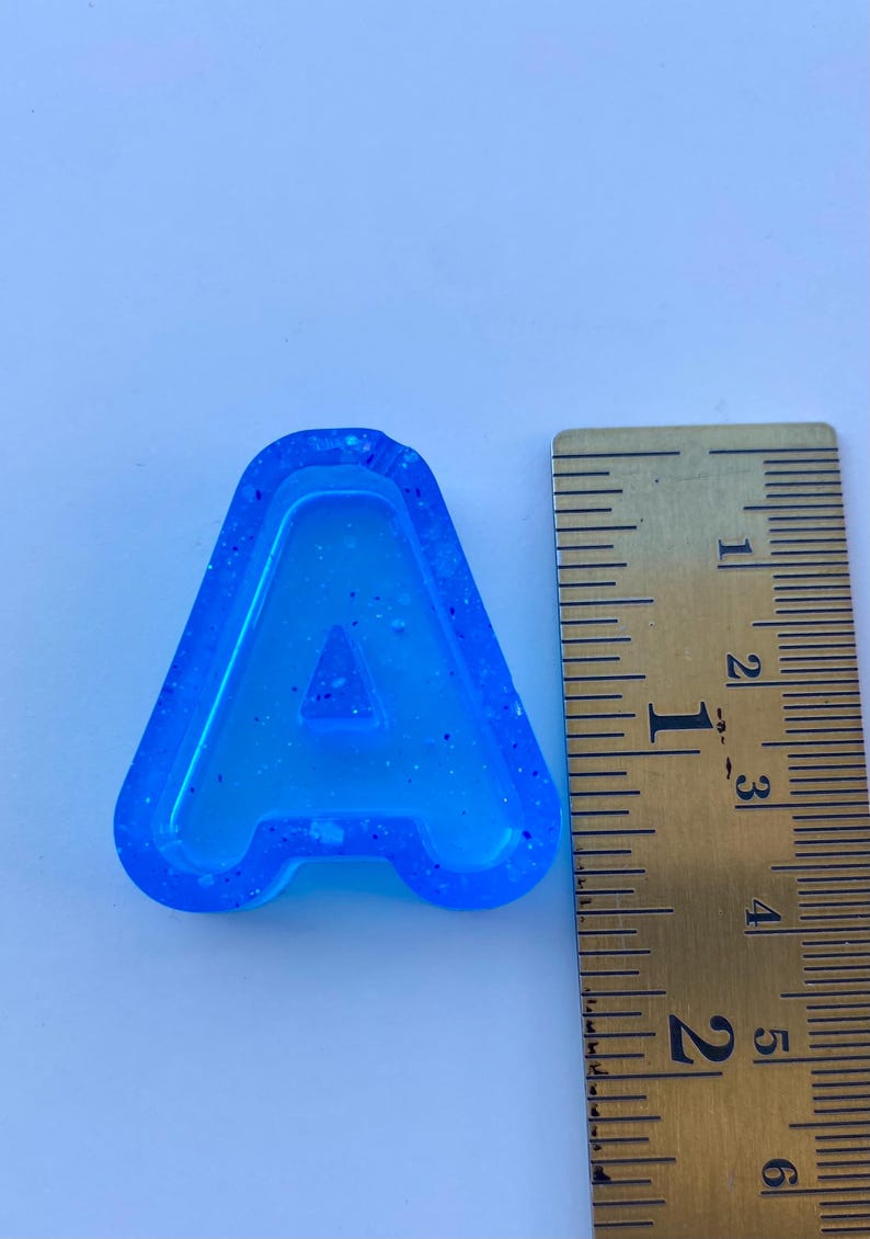 May include: A blue resin letter "A" with a glittery texture, next to a ruler. The letter is approximately 5 cm tall. The background is a light blue.