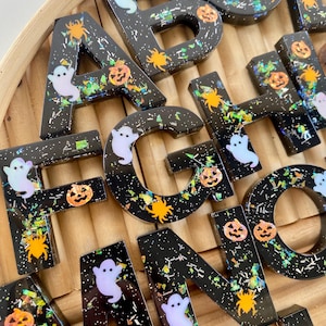 May include: A collection of black resin alphabet letters featuring embedded holographic glitter, orange spider shapes, and white ghost figures. Each letter has a glossy finish and contains various festive Halloween-themed confetti pieces.