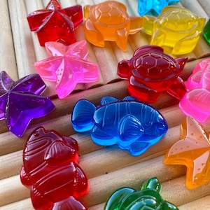 May include: A collection of colorful, translucent sea creature-shaped objects. The assortment includes starfish, turtles, and fish in various colors like red, orange, pink, purple, blue, yellow, and green. The objects are arranged on a light brown, wooden surface.