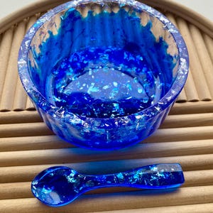 May include: A blue resin bowl and spoon set. The bowl is round with a textured interior and silver flecks. The spoon is also blue resin with silver flecks. The set is displayed on a wooden surface.