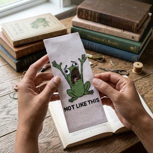 May include: A white bookmark with a cross-stitched green frog with its mouth open and arms raised. The text "NOT LIKE THIS" is below the frog. The background includes books with titles like "The Book of Frogs."