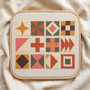 May include: A square embroidery hoop containing a piece of off-white fabric with twelve distinct cross-stitch squares arranged in a grid. Each square features geometric patterns in shades of mustard yellow, terracotta, olive green, and pink.