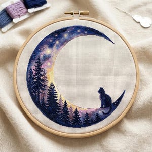 Könnte beinhalten: Ein runder Stickrahmen aus Holz mit einem Kreuzstichmotiv, das eine Mondsichel vor einem Sternenhimmel zeigt. Der Mond hat einen Farbverlauf in Gelb, Rosa und Dunkelblau, ergänzt durch Silhouetten von Tannenbäumen und einer sitzenden Katze.