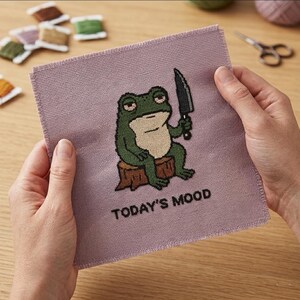 May include: A cross-stitch design on a lavender fabric square. The design features a green frog holding a knife, sitting on a tree stump, with the text "TODAY'S MOOD" below. The frog has a grumpy expression.