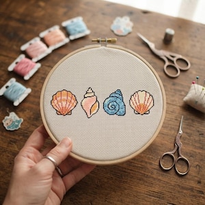 May include: A circular wooden embroidery hoop containing a piece of off-white fabric with a cross-stitch pattern of four seashells in shades of pink, orange, and blue, arranged horizontally across the center of the fabric.