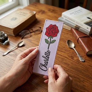 May include: A rectangular bookmark made of light purple fabric with a cross-stitched design of a red rose with green leaves and stem. The name Charlotte is embroidered in black cursive script below the rose, with stitched edges.