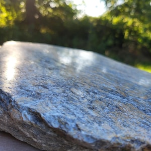 Może przedstawiać: Płaski, nieregularnie ukształtowany kawałek naturalnego kamienia o szorstkiej, teksturowanej powierzchni. Kamień ma nakrapianą paletę barw w odcieniach szarości, błękitu i ziemi, z lekko wypolerowaną górną powierzchnią odbijającą światło.