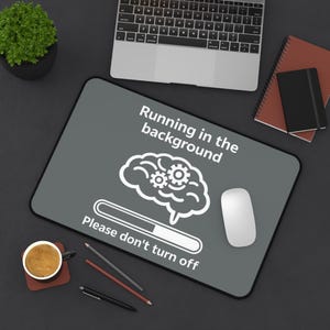 Può includere: Un tappetino per mouse grigio con il testo "Running in the background" e "Please don't turn off". Il tappetino presenta una grafica di un cervello bianco con ingranaggi e una barra di caricamento. Sono visibili anche un mouse bianco, un laptop e una tazza di caffè.