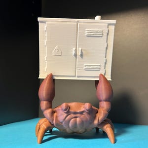 Puede incluir: Figura impresa en 3D de un cangrejo marrón y naranja sosteniendo un armario blanco. El armario tiene dos puertas y un pequeño símbolo. Las pinzas del cangrejo están levantadas.