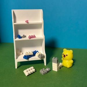 May include: A small white shelf unit with assorted toy building blocks in blue, pink, grey and white. A yellow bear figurine sits beside a small tower of blocks. The background is blue and green.