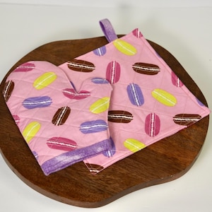 Puede incluir: Juego de manopla y agarradera rosas con un estampado de macarons de colores. Los macarons son amarillos, morados, rosas y marrones. El conjunto está sobre una tabla de madera. La agarradera tiene un lazo.