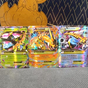 Op de afbeelding: Drie Pokémon-verzamelkaarten met Mega Venusaur, Mega Charizard en Mega Blastoise. De kaarten hebben kleurrijke illustraties en tekst, met een goudkleurige doos op de achtergrond. De kaarten liggen op een grijze ondergrond.