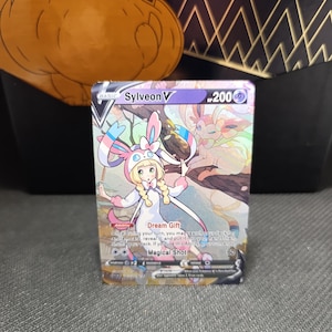 Puede incluir: Una tarjeta coleccionable Sylveon V con un personaje de dibujos animados con un atuendo blanco con detalles en rosa y azul. La tarjeta tiene el texto "Dream Gift" y "Magical Shot". El fondo incluye una caja dorada y negra.