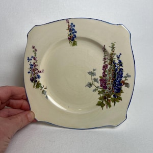 May include: Square, off-white plate with a blue rim and floral design. The plate features a central well and a decorative border. The floral design includes purple, blue, and green flowers.