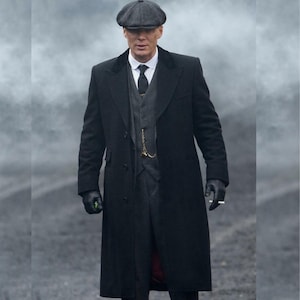 Puede incluir: Un hombre con un abrigo largo negro, chaleco gris y traje oscuro, con una gorra plana gris. Lleva una camisa blanca con cuello y una corbata negra. También lleva guantes negros y sostiene un cigarrillo. El fondo es una atmósfera gris y humeante.