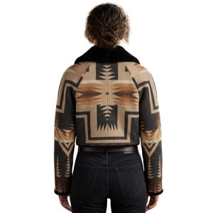 May include: A cropped jacket featuring a geometric pattern in shades of tan, brown, and charcoal gray. The garment has long sleeves, a black faux fur collar, and a straight hemline, worn over dark charcoal gray denim jeans with a brown leather belt.