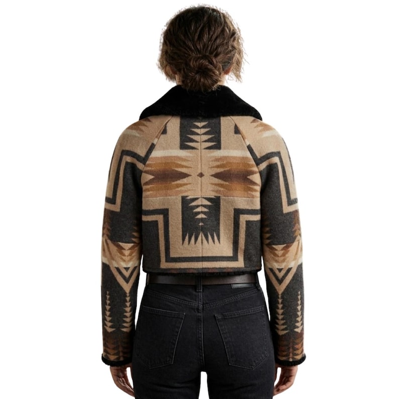 May include: A cropped jacket featuring a geometric pattern in shades of tan, brown, and charcoal gray. The garment has long sleeves, a black faux fur collar, and a straight hemline, worn over dark charcoal gray denim jeans with a brown leather belt.