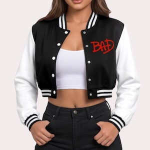 May include: Black and white cropped varsity jacket with the word "BAD" in red script on the chest. The jacket has white sleeves, a black body, and white and black striped cuffs and collar. It features a button-up front.
