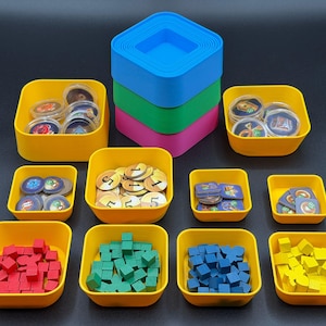 May include: A collection of square storage bowls in yellow, blue, green, and pink, containing various game components including small wooden cubes in red, green, blue, and yellow, circular tokens with numbers, and square tiles with graphic designs.