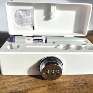 May include: A white, lockable case with a combination lock. Inside, a medication pen labelled "Once weekly" and a small packet are visible. The case is open, revealing the contents, and sits on a wooden surface.