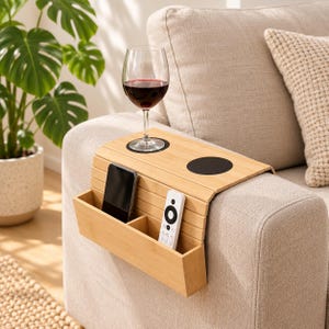 May include: A light-colored bamboo sofa armrest tray featuring a flexible slatted design, two circular black cup holders, and an attached storage compartment holding a black smartphone and a white remote control.