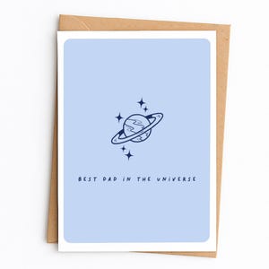 May include: A greeting card with a light blue background and a navy blue illustration of a planet with rings and stars. The text "BEST DAD IN THE UNIVERSE" is printed in navy blue at the bottom of the card. The card is inside a brown envelope.
