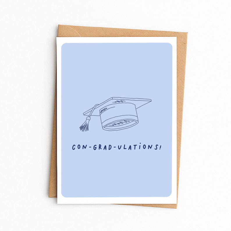 Op de afbeelding: Een lichtblauwe wenskaart met een donkerblauwe lijntekening van een afstudeerhoed met een kwastje. Onder de hoed staat de tekst CON-GRAD-ULATIONS! in donkerblauwe hoofdletters gedrukt. De kaart ligt op een bruine papieren envelop.