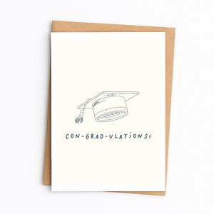 Op de afbeelding: Een cr&egrave;mekleurige wenskaart met een eenvoudige zwarte lijntekening van een afstudeerhoed met een kwastje. Onder de illustratie staat de tekst CON-GRAD-ULATIONS! in het zwart gedrukt. De kaart zit in een bruine envelop van kraftpapier.