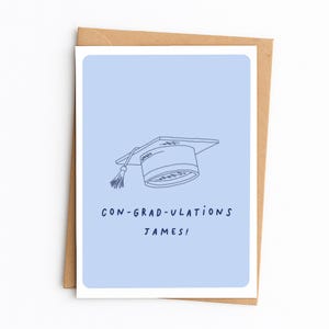 Op de afbeelding: Een lichtblauwe wenskaart met een donkerblauwe lijntekening van een afstudeerhoed met een kwastje. Onder de illustratie staat de tekst CON-GRAD-ULATIONS JAMES! in donkerblauwe hoofdletters. De kaart ligt op een bruine envelop.