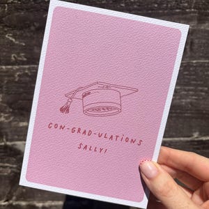 Op de afbeelding: Een rechthoekige wenskaart met een getextureerde mauve achtergrond met een donkerrode lijntekening van een afstudeerhoed met een kwastje. De tekst in donkerrood onder de hoed luidt CON-GRAD-ULATIONS SALLY! De kaart heeft een witte rand.