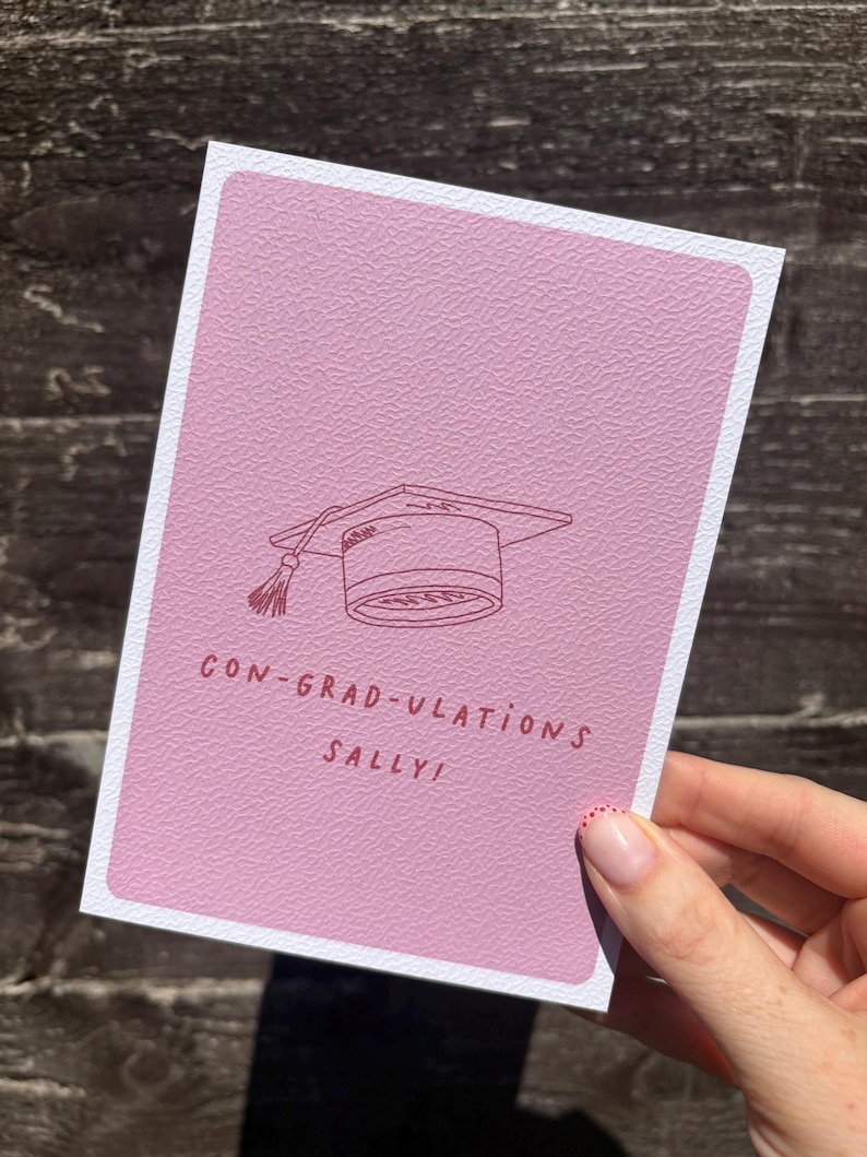 Op de afbeelding: Een rechthoekige wenskaart met een getextureerde mauve achtergrond met een donkerrode lijntekening van een afstudeerhoed met een kwastje. De tekst in donkerrood onder de hoed luidt CON-GRAD-ULATIONS SALLY! De kaart heeft een witte rand.