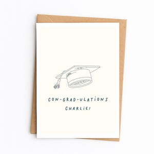 Op de afbeelding: Een witte wenskaart met een donkerblauwe lijntekening van een afstudeerhoed met een kwastje. Onder de illustratie staat de tekst CON-GRAD-ULATIONS CHARLIE! in donkerblauwe hoofdletters. De kaart ligt op een bruine envelop.