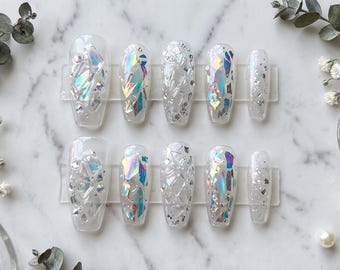 Puede incluir: Un conjunto de uñas postizas transparentes e iridiscentes con una variedad de diseños. Algunas uñas presentan grandes fragmentos iridiscentes coloridos, mientras que otras tienen un efecto de purpurina plateada. Las uñas están dispuestas sobre una superficie de mármol blanco.