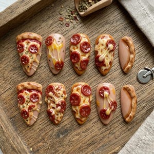 Puede incluir: Una colección de diseños de uñas con temática de pizza. Cada uña presenta una porción de pizza en miniatura con pepperoni, queso y corteza. Algunas uñas tienen queso goteando. Las uñas están dispuestas en una bandeja de madera.