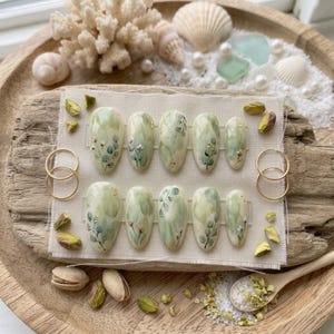 May include: A set of ten almond-shaped press-on nails featuring a soft sage green watercolor wash, delicate hand-painted botanical eucalyptus leaf motifs, and tiny gold foil accents. The nails are arranged in two rows on a rectangular fabric piece.