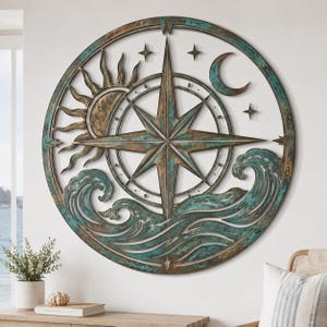 May include: A circular wall art piece featuring a central compass rose, a sun, a crescent moon, and stylized ocean waves. The design is finished in a weathered bronze and teal patina, with small four-pointed stars scattered across the frame.