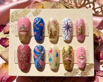 Peut inclure: Un ensemble de dix faux ongles avec divers motifs. Les ongles présentent des couleurs rose, bleu, or et blanc, avec des motifs comprenant des couronnes, des roses, des châteaux et des étoiles. Certains ongles ont des accents de paillettes et des perles. Les ongles sont disposés sur un présentoir en bois.