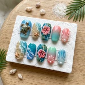 May include: A set of ten press-on nails with a beach theme. Designs include a sea turtle, starfish, hibiscus flowers, and seashell patterns. Colours include blue, tan, pink, and green. The nails are arranged on a white rectangular surface.