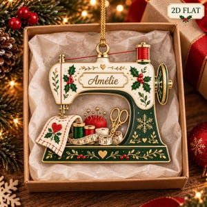 May include: A flat, cream and dark green sewing machine ornament featuring the name Amélie, decorated with holly berries, leaves, and gold accents. It includes a red pincushion, gold scissors, thread spools, and a measuring tape.