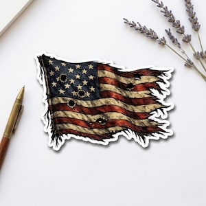 May include: A sticker featuring a distressed American flag with a tattered edge and multiple dark bullet hole graphics. The flag displays the traditional red, white, and blue color scheme with a weathered, aged appearance and visible ink-style lines.