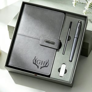 Peut inclure: Un carnet gris avec une fermeture à sangle magnétique et un logo en forme de bois de cerf gravé avec les lettres GH. L'ensemble comprend un stylo à bille gris assorti, une recharge fine en métal et une clé USB rectangulaire dans une boîte grise.